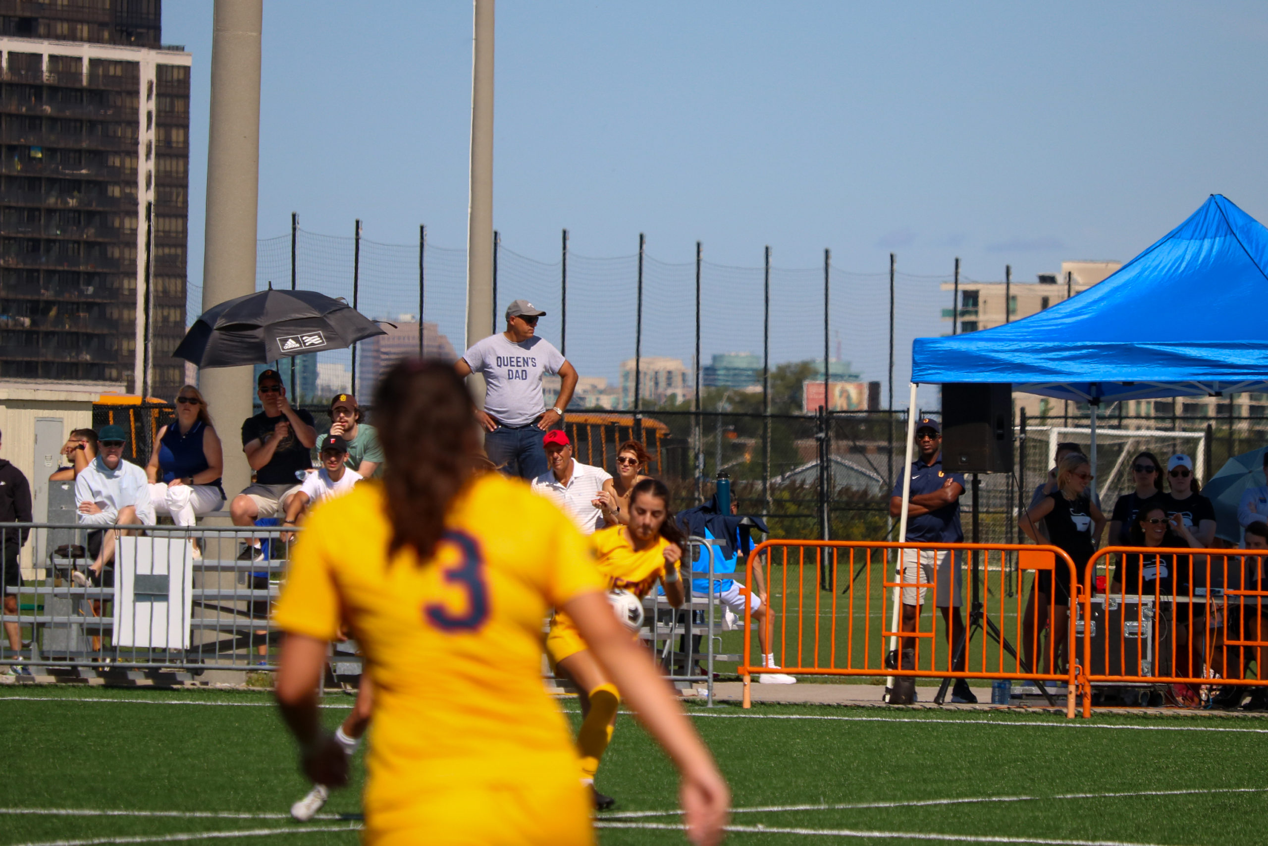 In the stands behind soccer players in the middle of a game is a man in a grey "Queen's dad" shirt