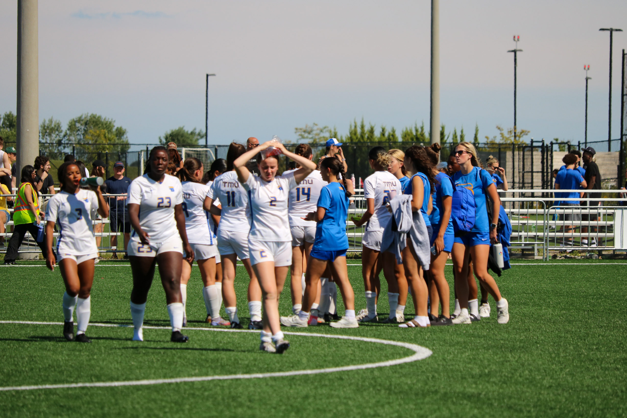 TMU Bold women's soccer players in white jerseys (and some in blue t shirts) gather after a game. One player has her hands on her head