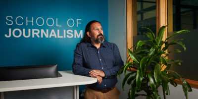 A man with long dark hair and a beard stares in the distance leaning against a reception desk.