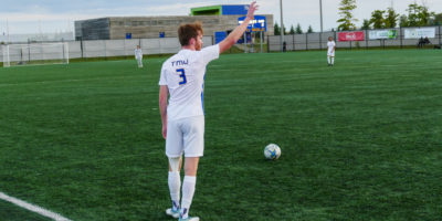 TMU Bold soccer player COlin Gander prepares to take a free kick on the soccer field at Downsview Park. He s wearing a white jersey and there is a blue and gold scoreboard visible in the background