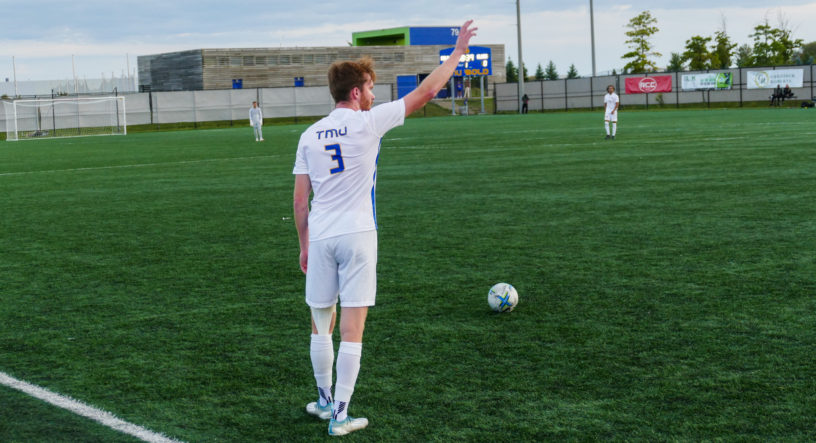 TMU Bold soccer player COlin Gander prepares to take a free kick on the soccer field at Downsview Park. He s wearing a white jersey and there is a blue and gold scoreboard visible in the background