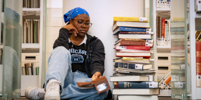 An upset student stares at their wallet sitting on a library floor, next to a stack of books