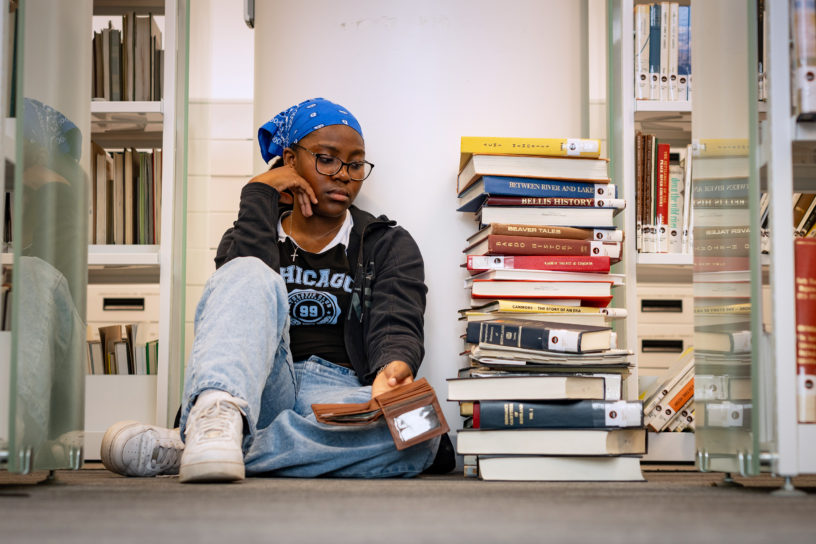 An upset student stares at their wallet sitting on a library floor, next to a stack of books