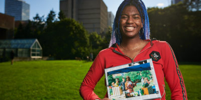 A student stands in the field, smiling at the camera holding an old photo of herself