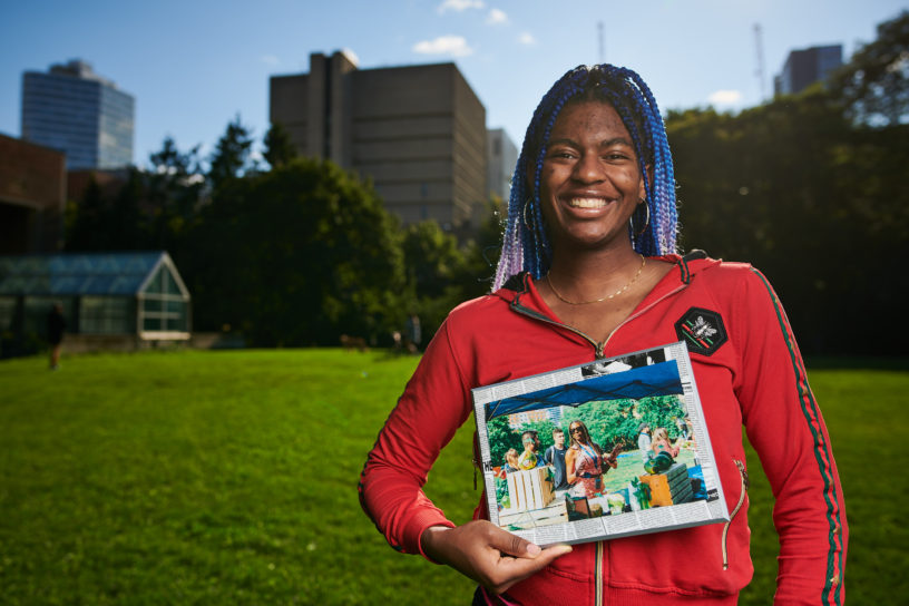 A student stands in the field, smiling at the camera holding an old photo of herself