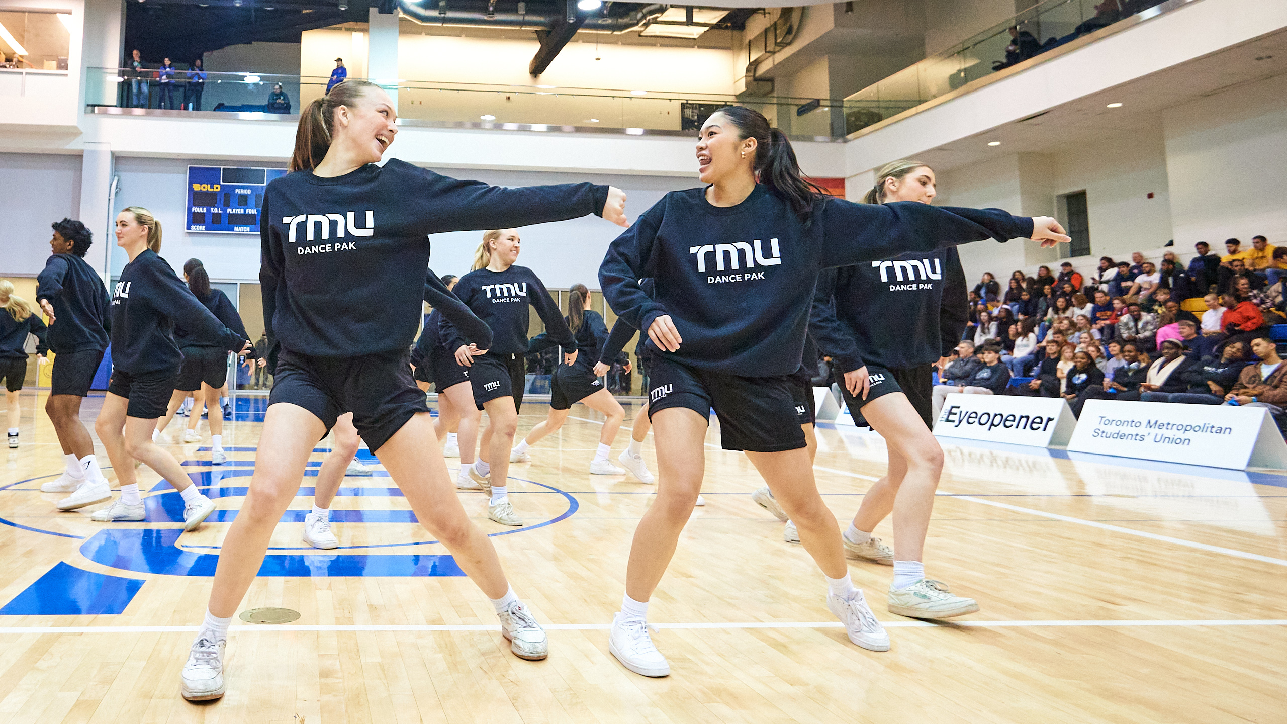 A group of students in black 'TMU' sweaters perform a dance number in a gymnasium