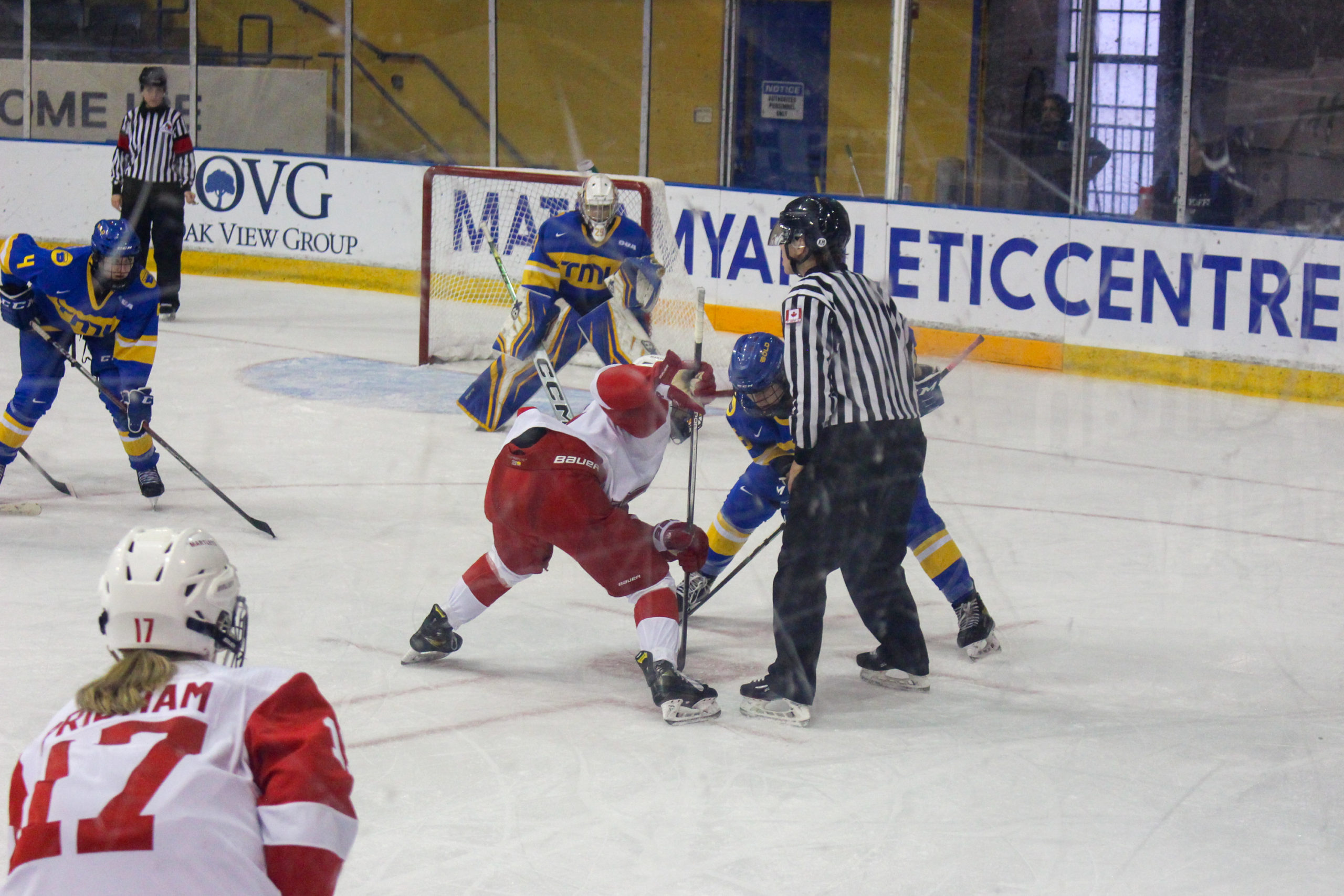 A TMU Bold player and a McGill Martlets player take a faceoff in front of TMU goalie Alexia Stratos