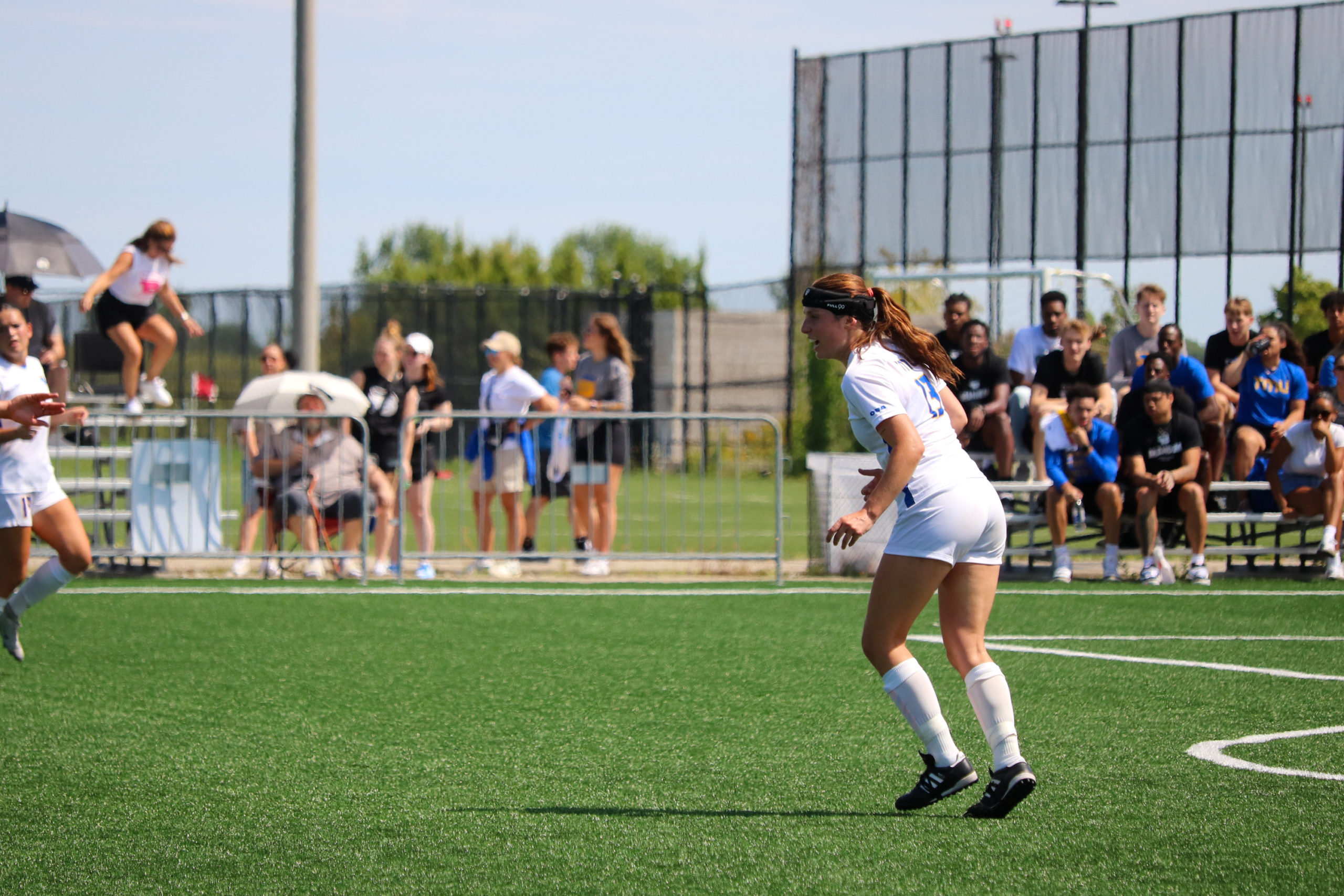 A player in a white TMU jersey with the number 13 prepares to kick a soccer ball. Fans in blue and yellow can be seen in the background..