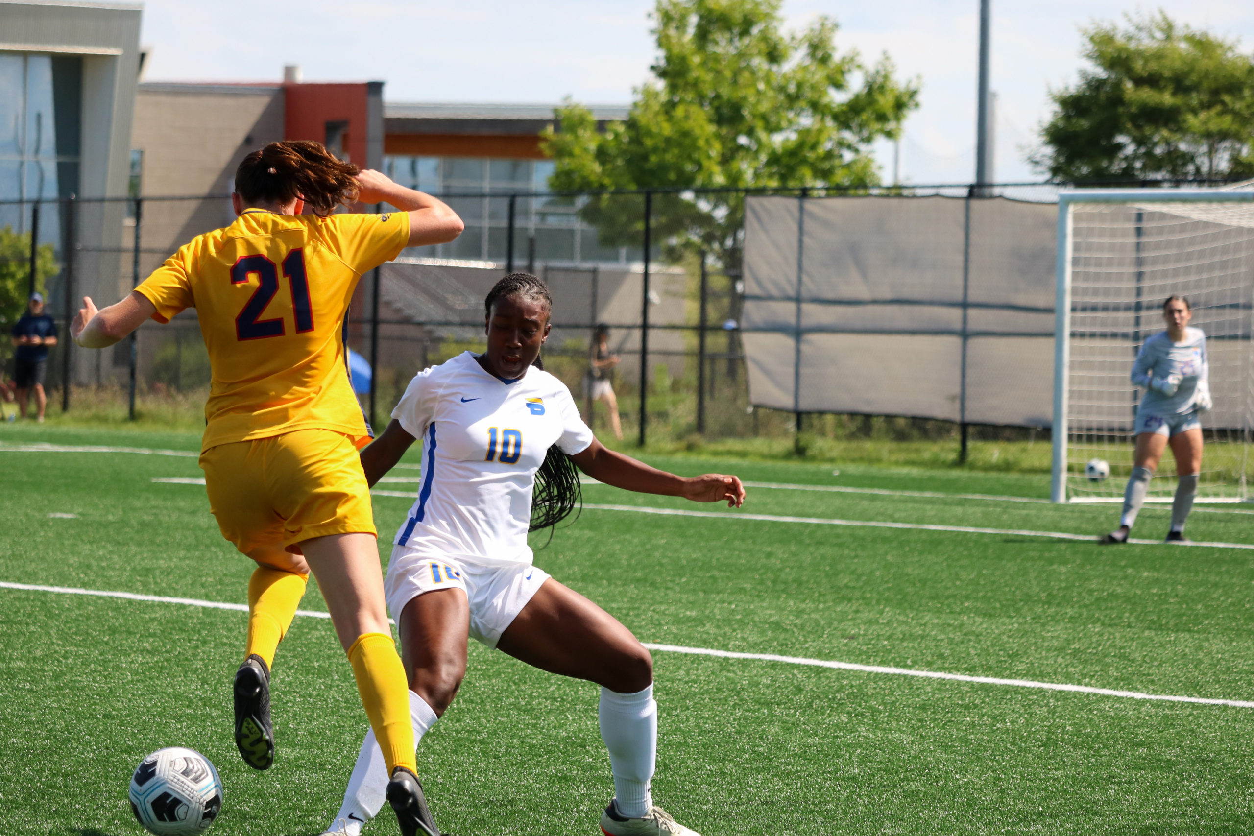 A women's soccer player in a white uniform reaches her foot  to kock a soccer ball away from the feet of a player in a gold uniform.