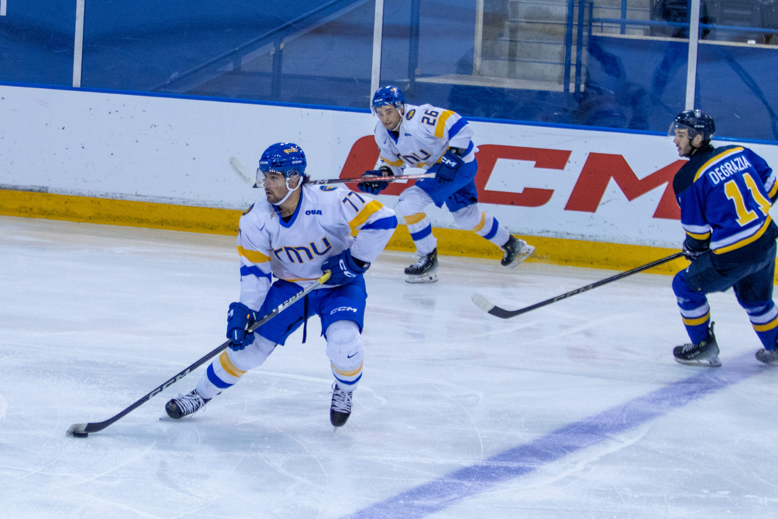 Two TMU Bold men's hockey players in white jerseys prepare to head up the ice with the puck while a player in a blue Lakehead jersey lurks.