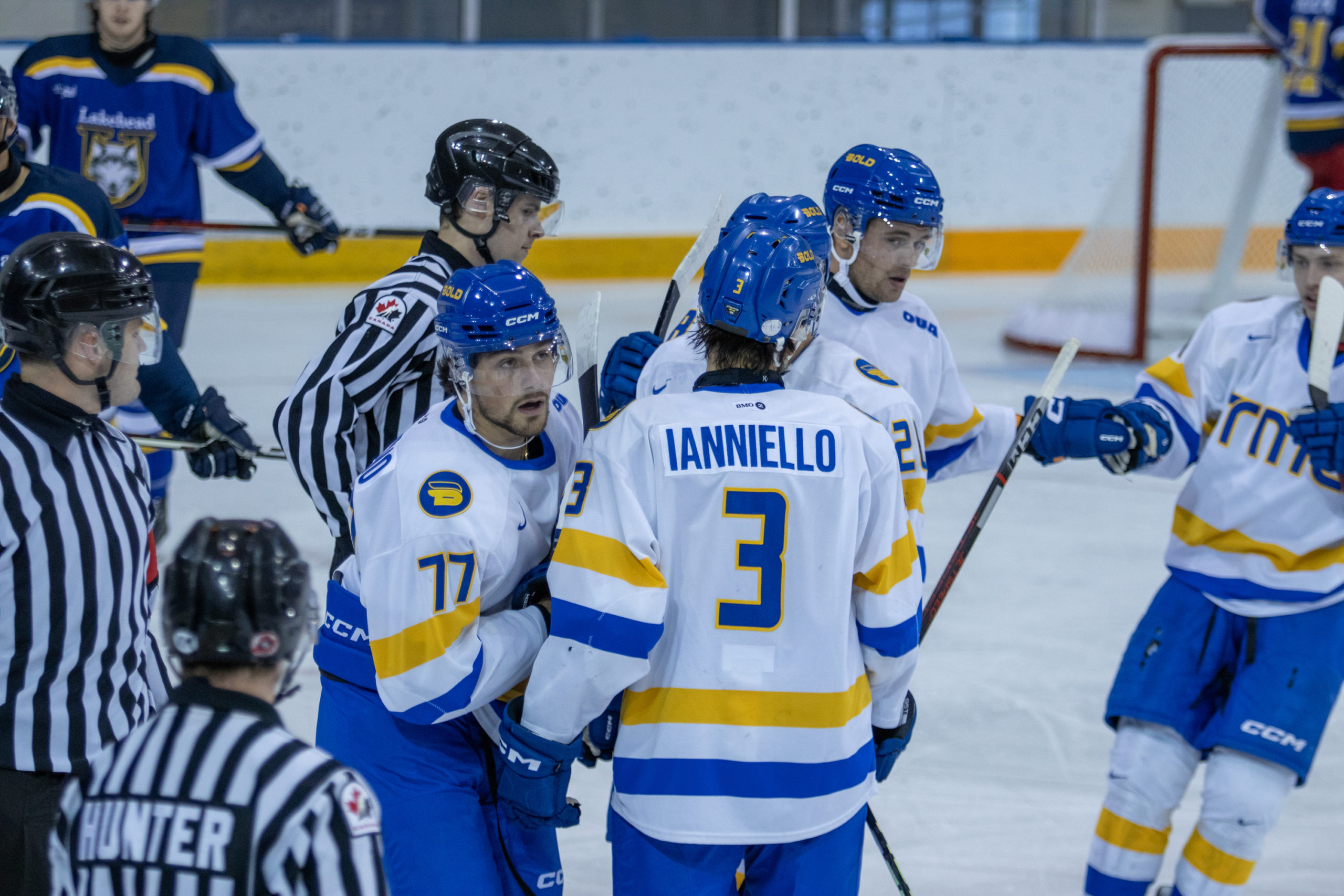 TMU Bold men's hockey players in white jerseys gather around their teammate to celebrate a goal. Two referees in striped uniforms are in the picture.