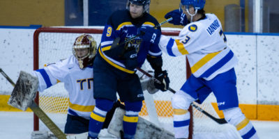 A TMU Bold men's hockey player in a white jersey cross-checks a player in a blue and black Lakehead Thunderwoles uniform in front of a TMU goaltender