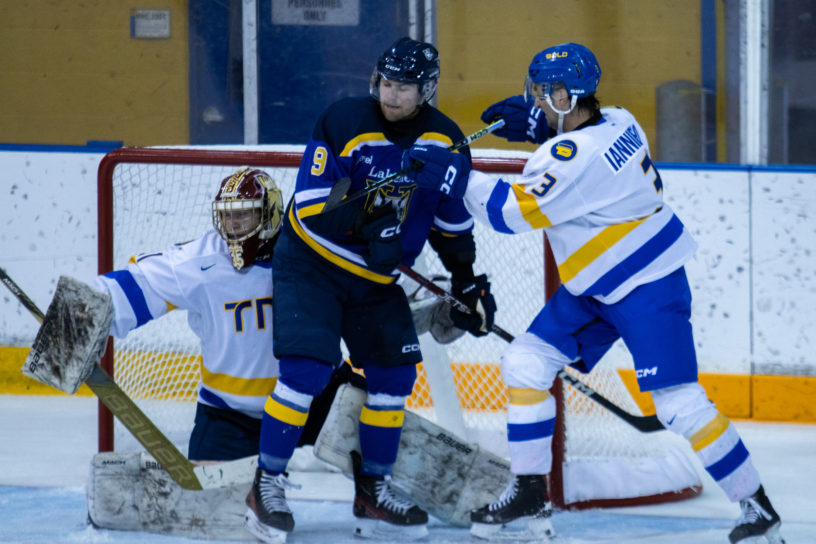 A TMU Bold men's hockey player in a white jersey cross-checks a player in a blue and black Lakehead Thunderwoles uniform in front of a TMU goaltender