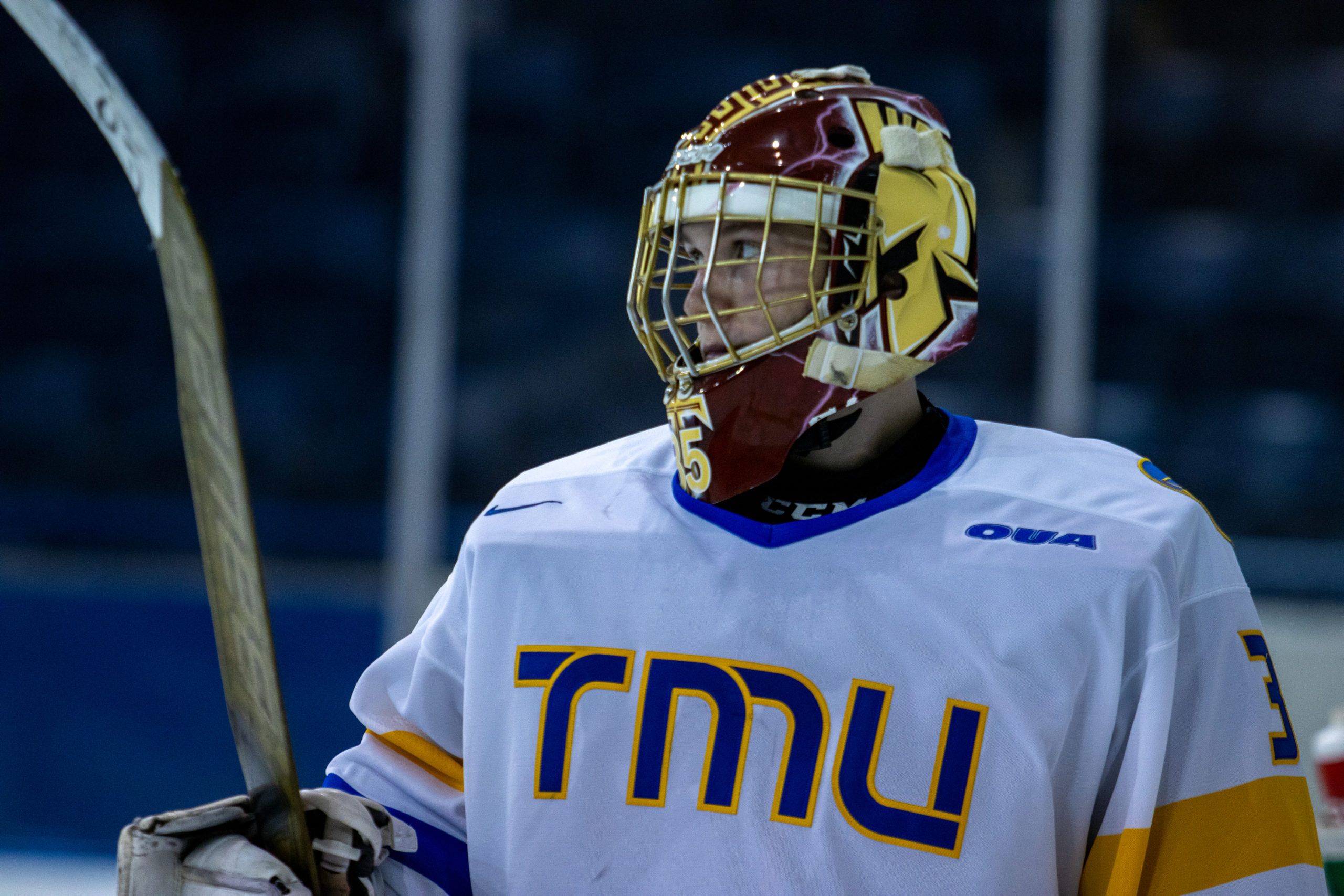 A close-up photo shows a goaltender in a white TMU Bold jersey and wearing a maroon and gold mask. He has his stick blade in the air.