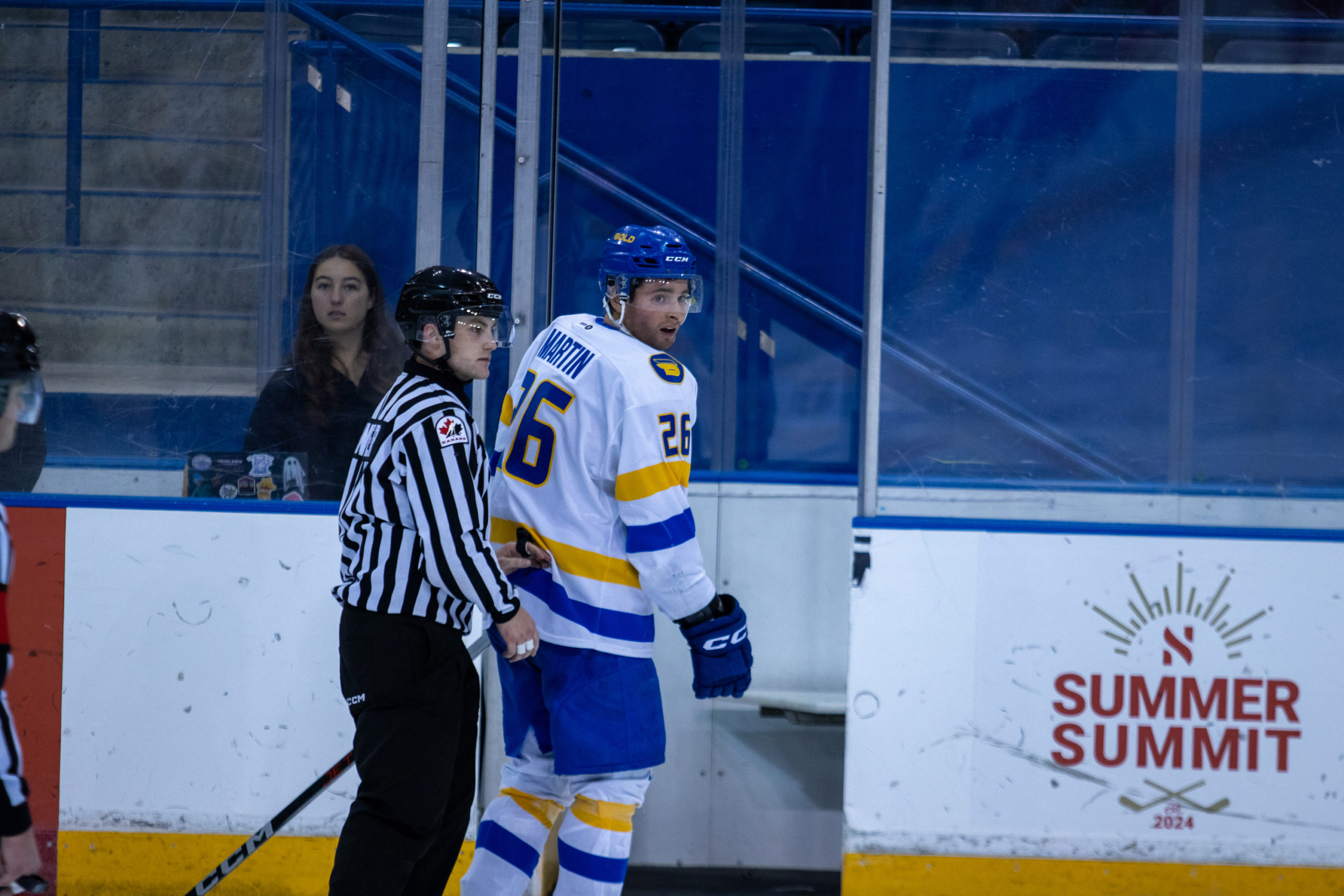 A men's hockey player in a white TMU Bold jersey looks backwards past the linesperson leading him to the penalty box with a questioning expression.