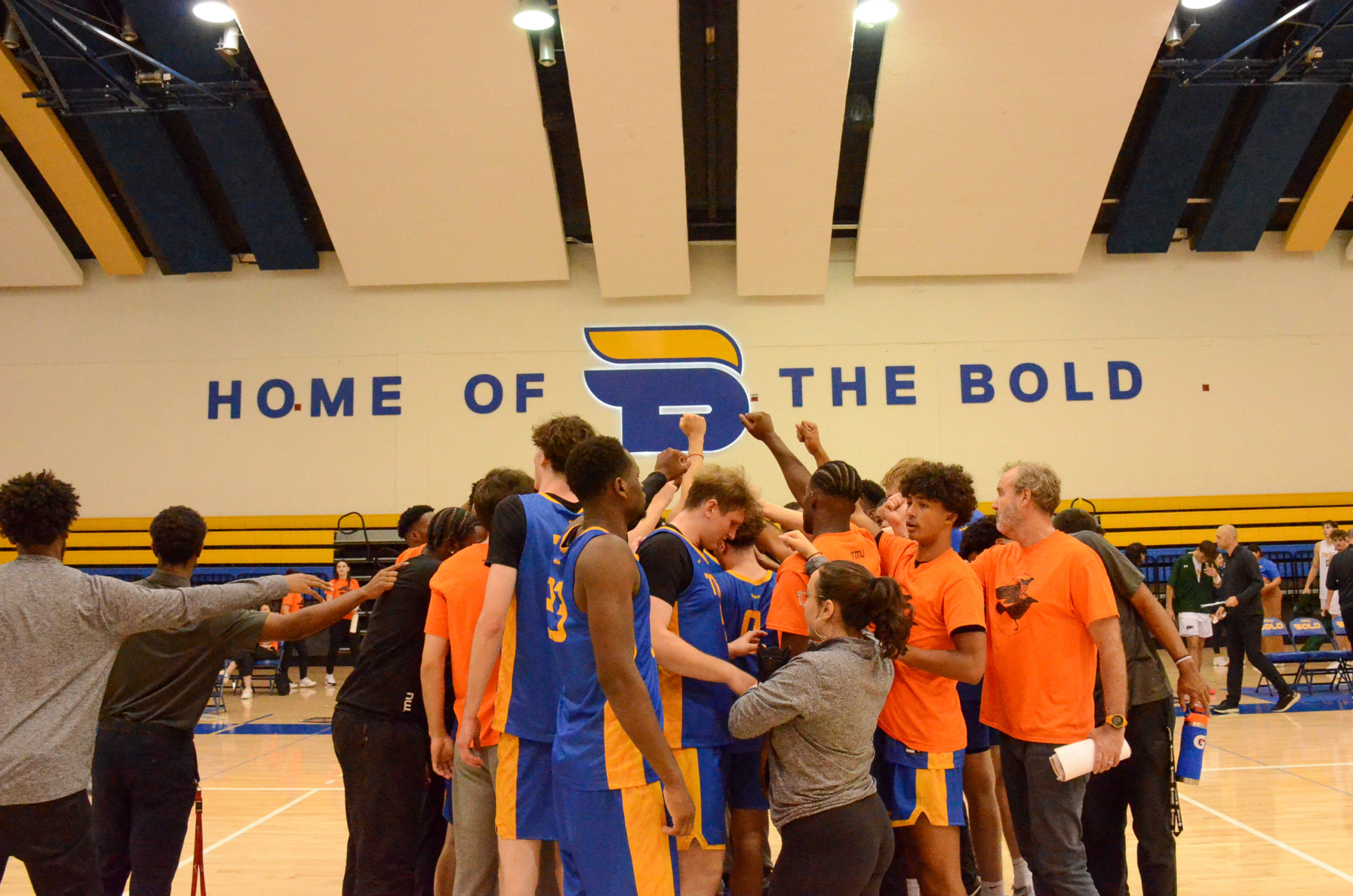 The TMU Bold men's basketball team gather at centre court and raise their fists in the air