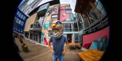 A man wearing a bird head poses in front of a mural.