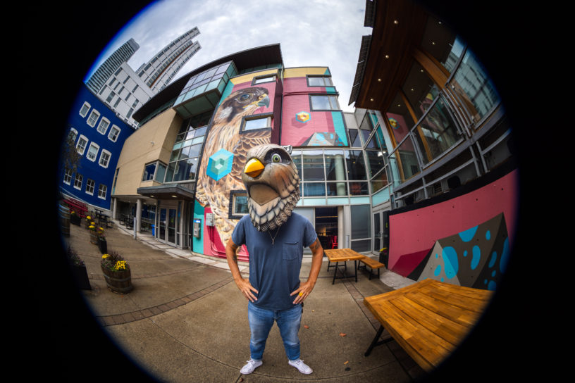 A man wearing a bird head poses in front of a mural.