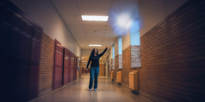 A young woman wearing a long-sleeved black shirt and blue jeans is in the middle of a long winding hallway surrounded by red lockers and brick walls. She is looking and reaching up toward a shiny white sphere of light floating above her head.