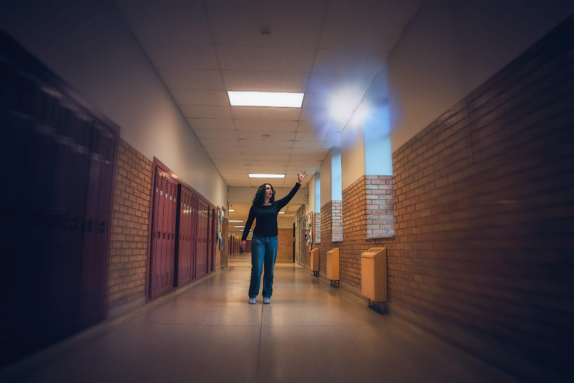 A young woman wearing a long-sleeved black shirt and blue jeans is in the middle of a long winding hallway surrounded by red lockers and brick walls. She is looking and reaching up toward a shiny white sphere of light floating above her head.