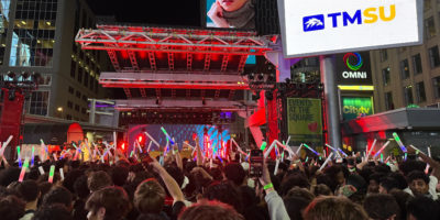 An audience of students stand crowded in front of a stage in preparation for a concert.