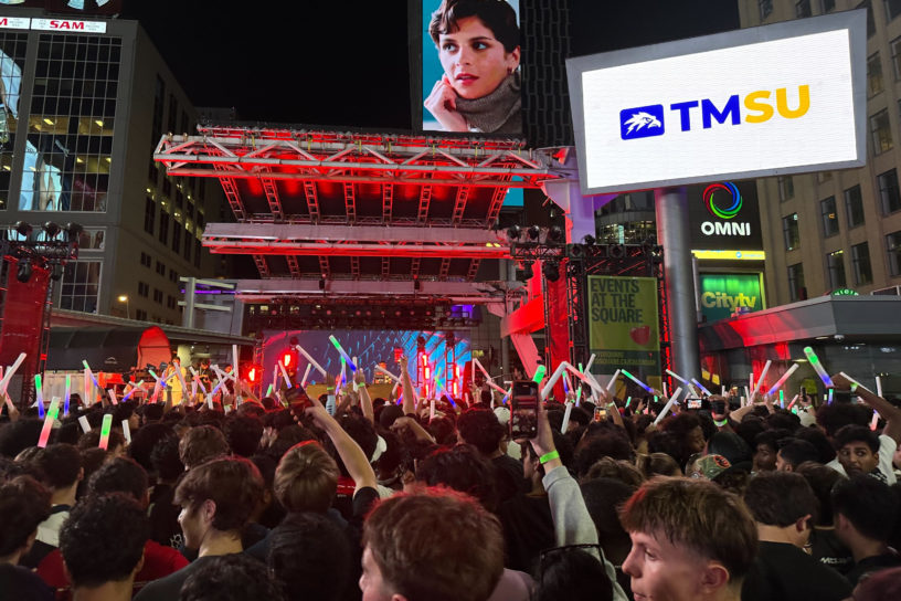 An audience of students stand crowded in front of a stage in preparation for a concert.