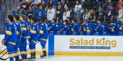 TMU Bold men's hockey players stand for the national anthem on the bench and at the blue line. fans stand behind the bench