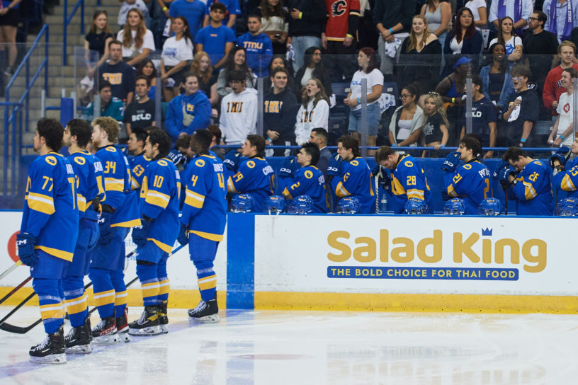 TMU Bold men's hockey players stand for the national anthem on the bench and at the blue line. fans stand behind the bench