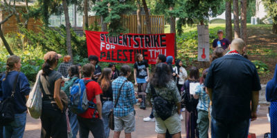 Students at Kerr Hall stand around red banner with "student strike for palestine TMU" written on it. A man standing next to the sign speaks with a megaphone