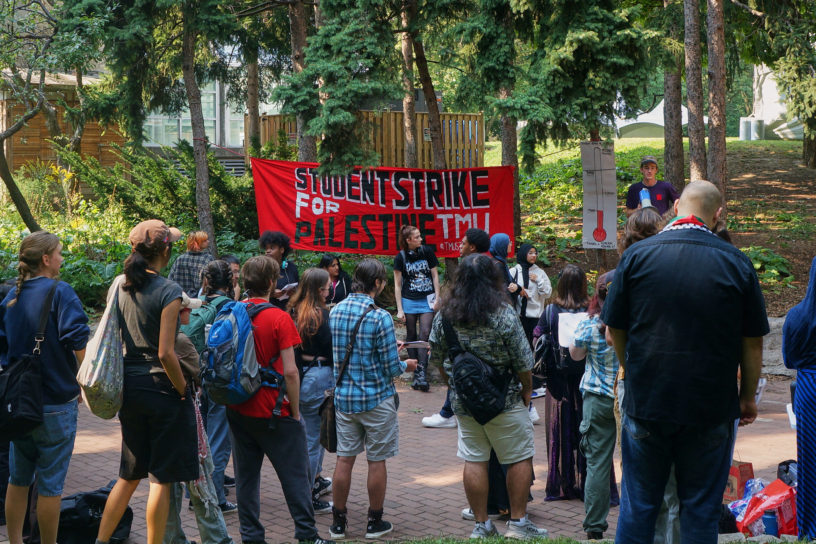 Students at Kerr Hall stand around red banner with "student strike for palestine TMU" written on it. A man standing next to the sign speaks with a megaphone