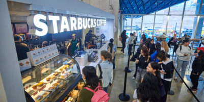 A side shot of a Starbucks coffee shop within a university building. A long line of students stand waiting to order.