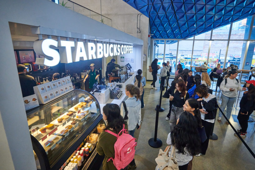 A side shot of a Starbucks coffee shop within a university building. A long line of students stand waiting to order.