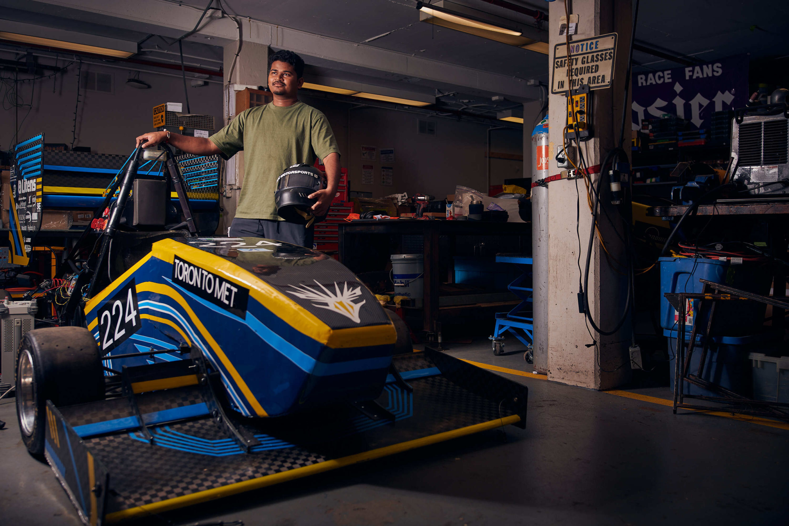 Rhythm Patel stands beside the blue and yellow TM-24 car in an empty workshop. The car features a TMFR logo on its hood.