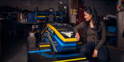 Rochelle Mendonca sits beside and looks at a blue and yellow electric race car in a shop.