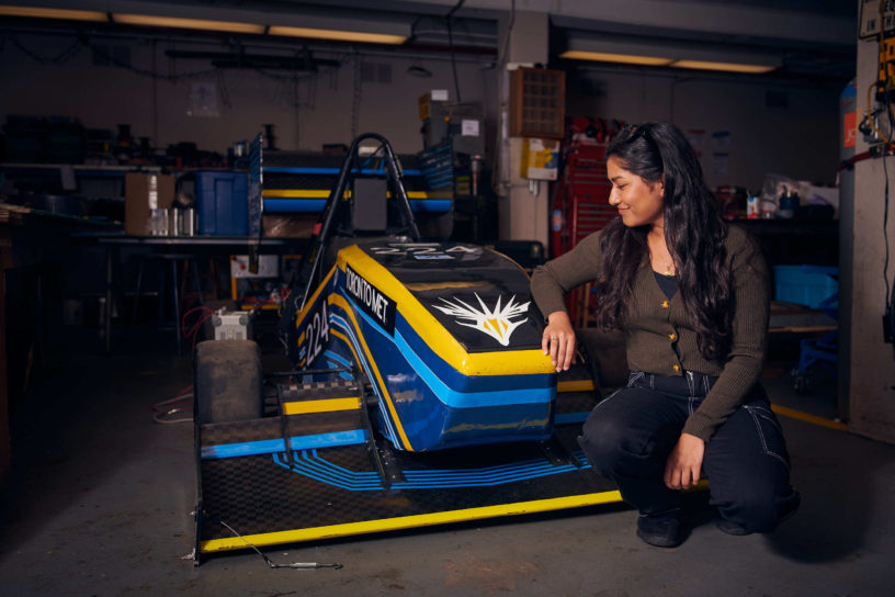 Rochelle Mendonca sits beside and looks at a blue and yellow electric race car in a shop.