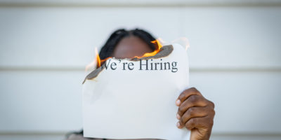 A person holds a burning piece of paper, with the words "We're Hiring" written on it.