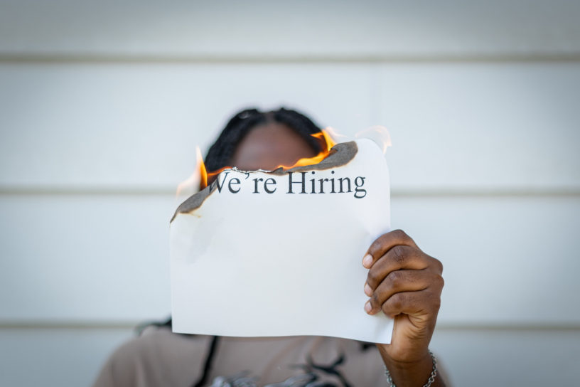 A person holds a burning piece of paper, with the words "We're Hiring" written on it.