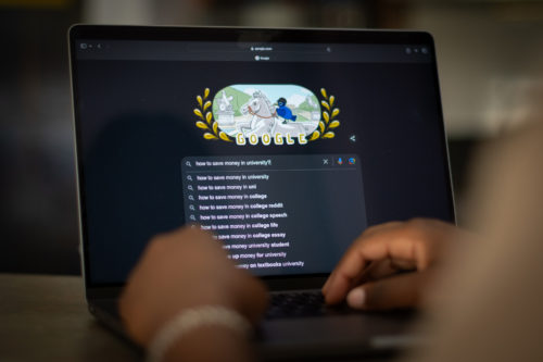 A close of shot of a person typing on a laptop searching up "how to save money in university?" on Google.