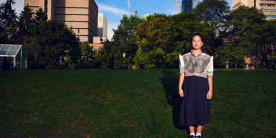 A student stands in the middle of an open field in the city, wearing a traditional filipiniana dress.