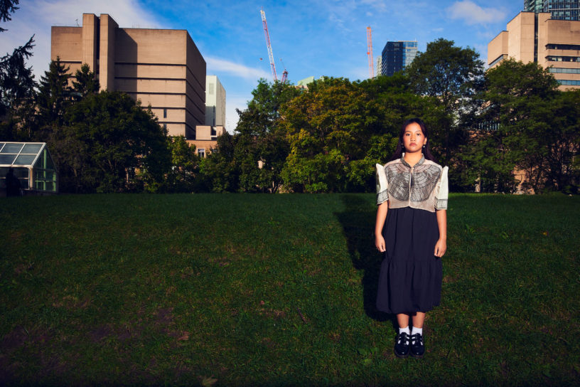 A student stands in the middle of an open field in the city, wearing a traditional filipiniana dress.