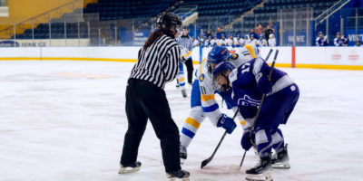 Hockey players in a white jersey and a purple jersey take a faceoff as a referee drops a puck