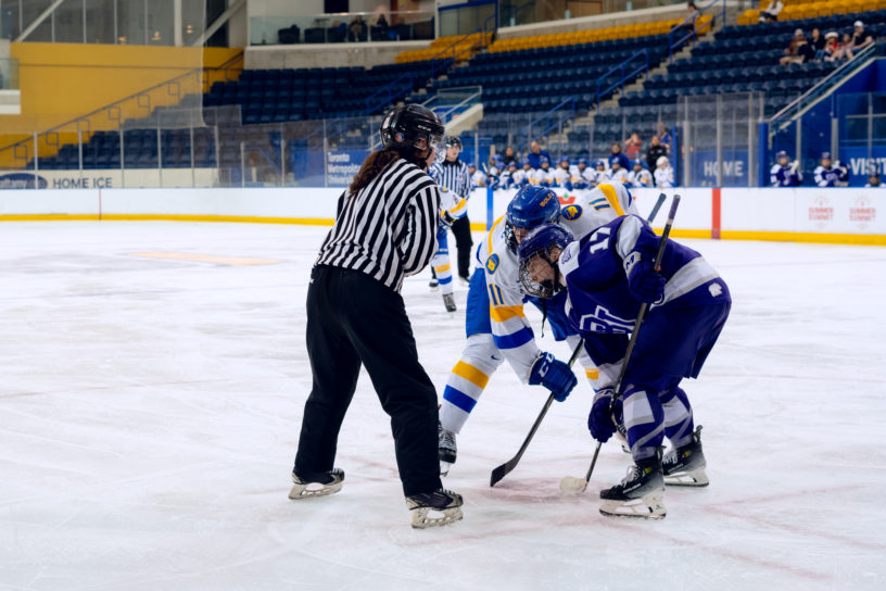 Hockey players in a white jersey and a purple jersey take a faceoff as a referee drops a puck