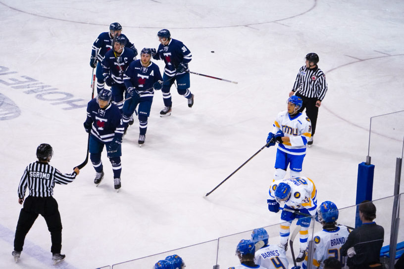 U of T Varsity Blues men's hockey players skate to their bench in a line after scoring, while TMU players stand around