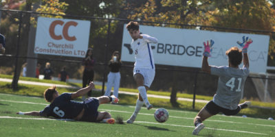 Justin Santos prepares to shoot a soccer ball past a goaltender jumping at him and a defender sliding behind him