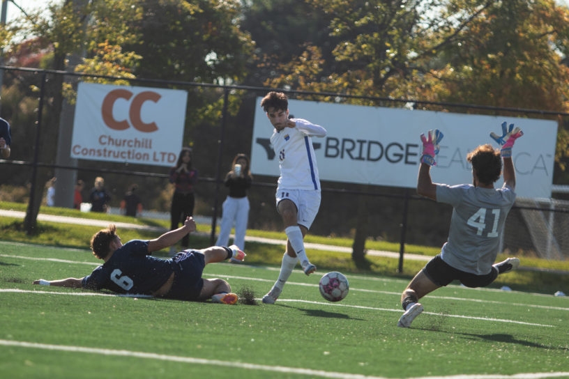 Justin Santos prepares to shoot a soccer ball past a goaltender jumping at him and a defender sliding behind him