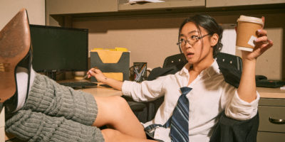 An office worker sits at a desk with her heels propped up, holding a cup of coffee and clicking her keyboard with a pen.