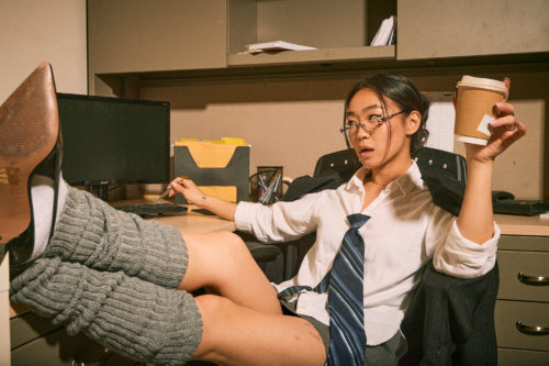 An office worker sits at a desk with her heels propped up, holding a cup of coffee and clicking her keyboard with a pen.