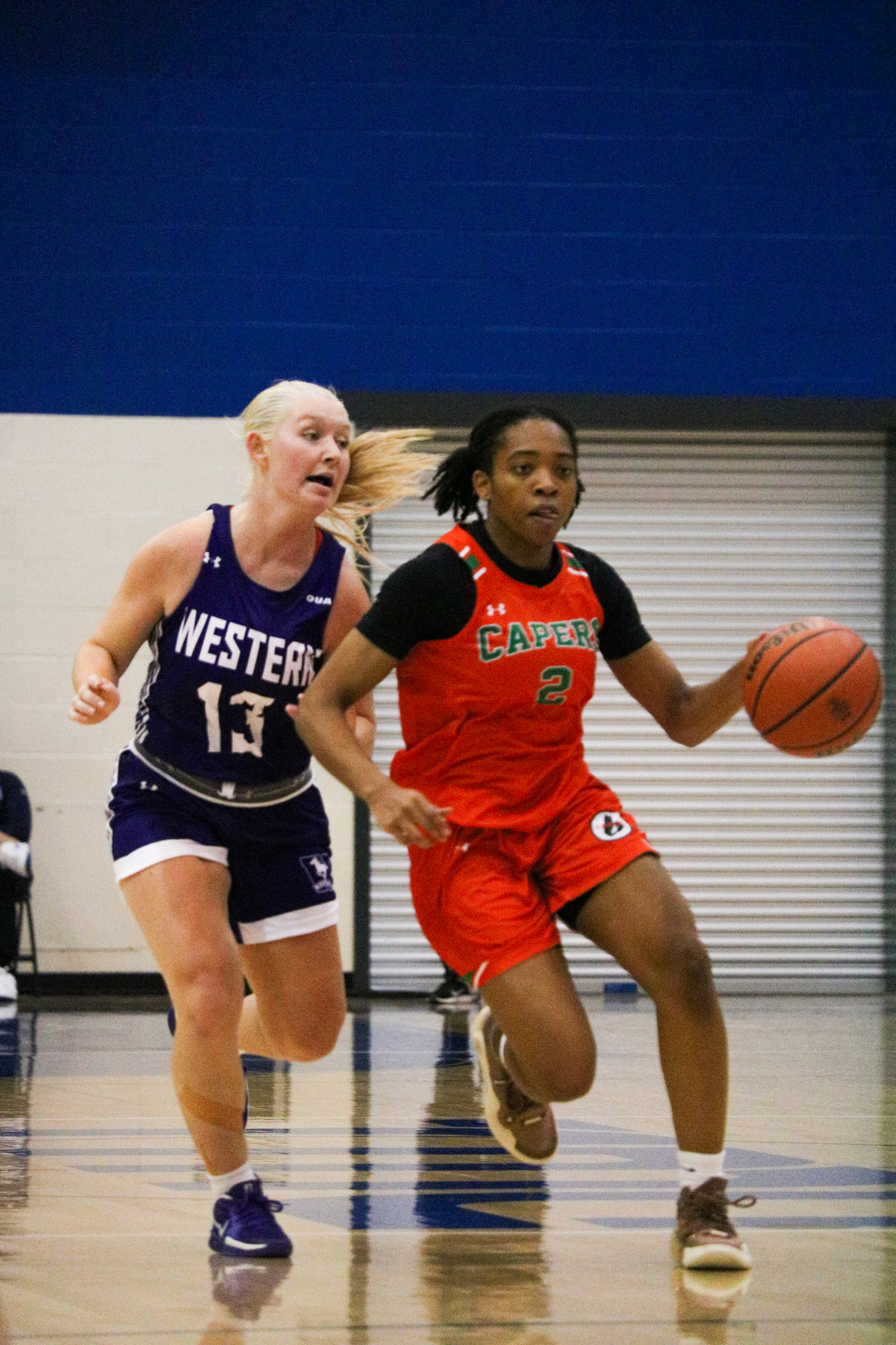 A Cape Breton Capers player dribbles a basketball as a Western Mustangs player chases her