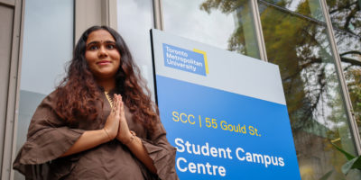 Woman outside of building with hands clasped in a 'Namaste' pose smiling and looking beyond the camera