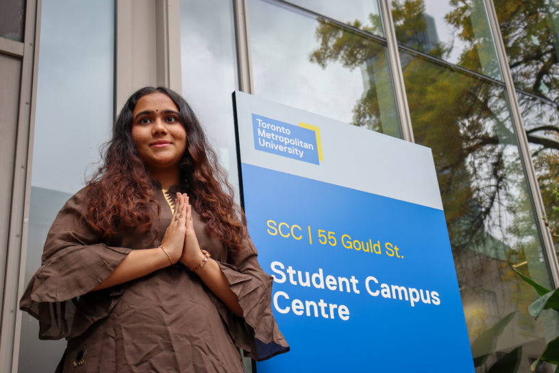 Woman outside of building with hands clasped in a 'Namaste' pose smiling and looking beyond the camera